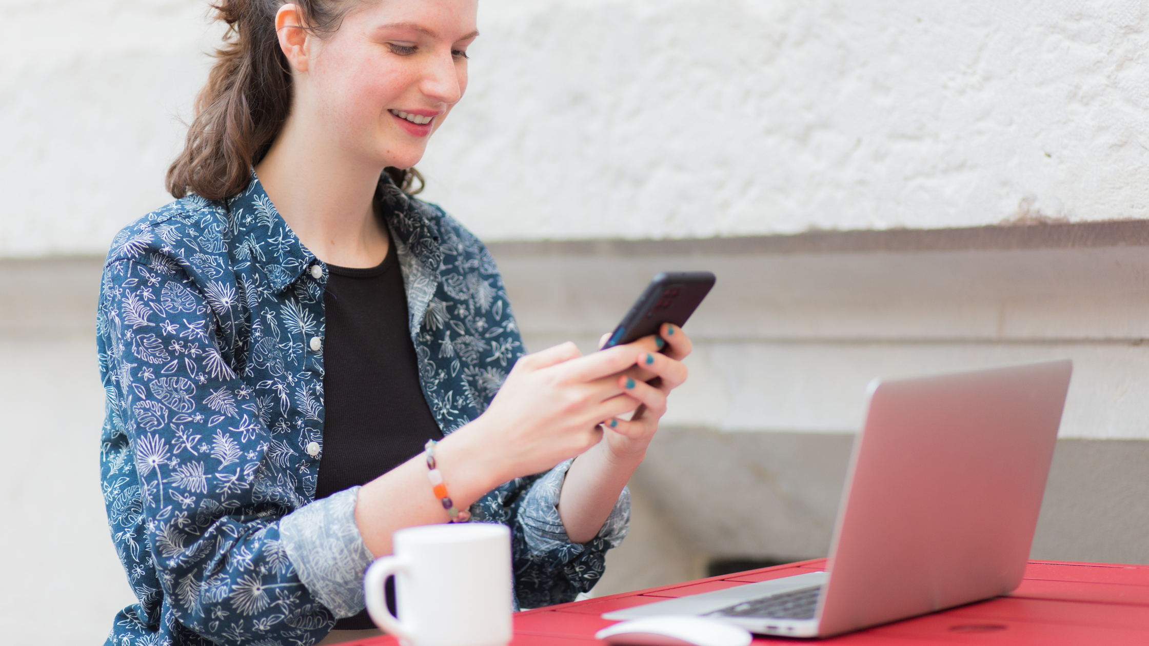 Woman sitting at a table using her phone with an open laptop in front of her