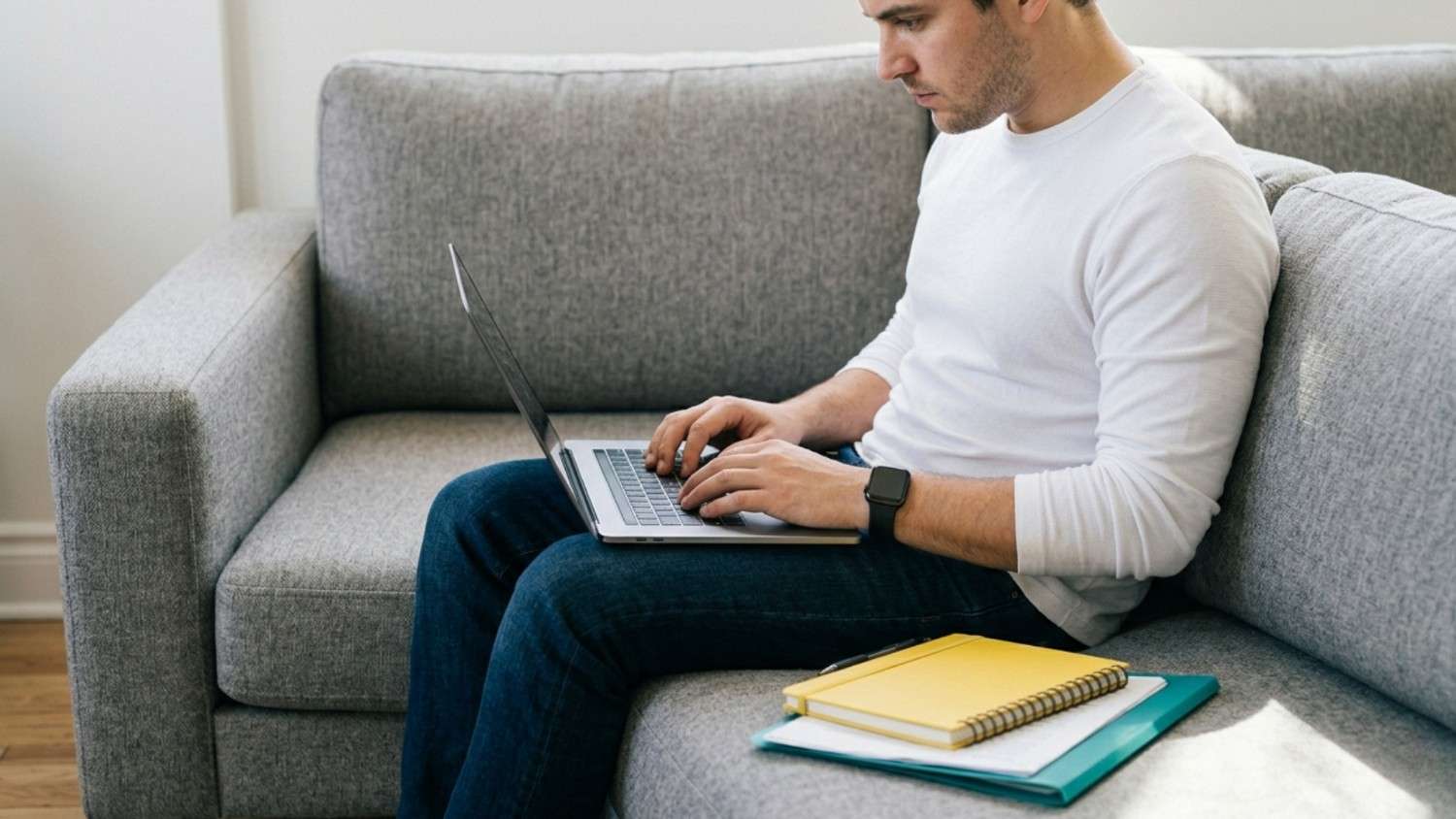 Man sitting on a gray couch using a laptop with notebooks beside him.